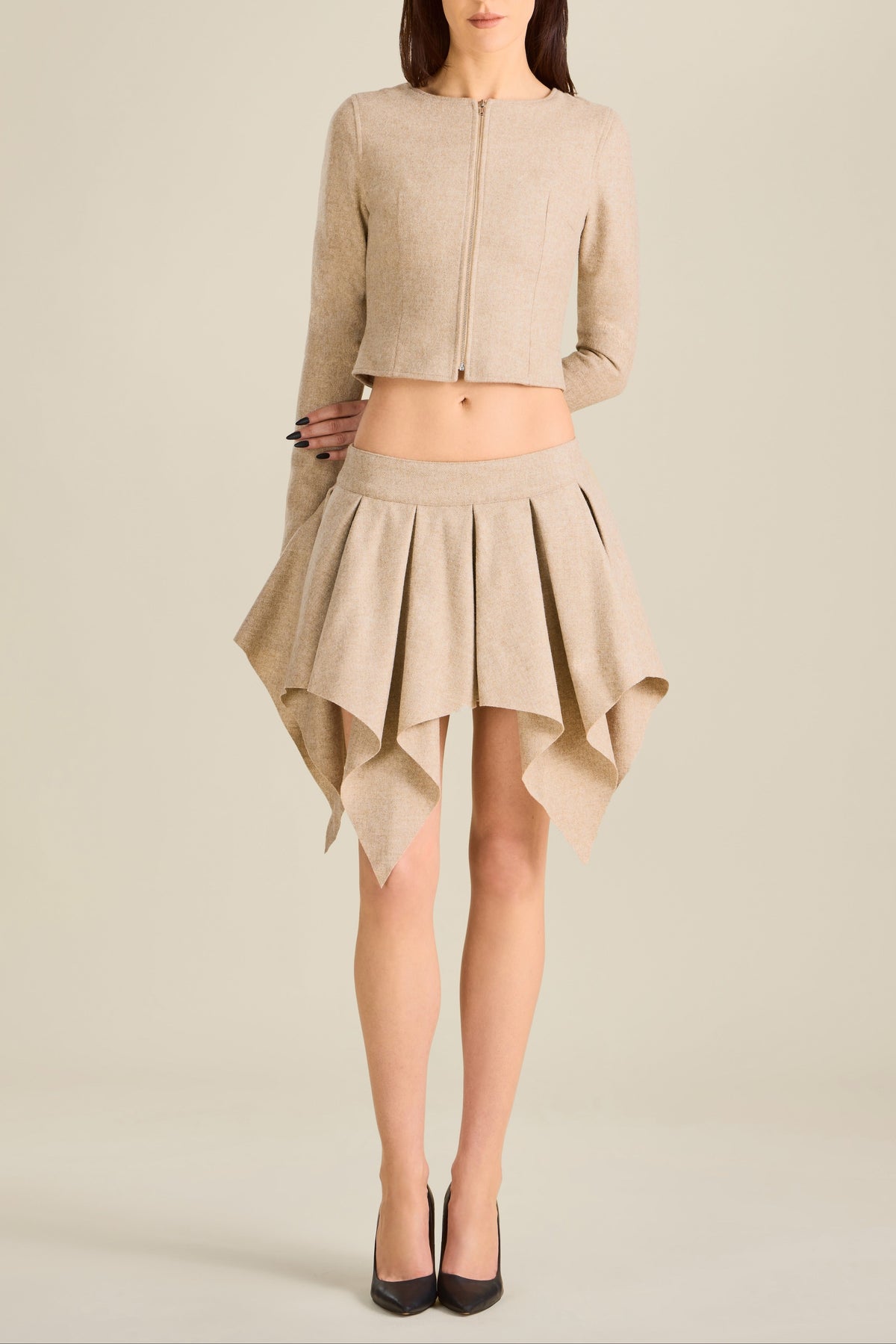Woman wearing a beige outfit with a jacket and pleated skirt on a plain background
